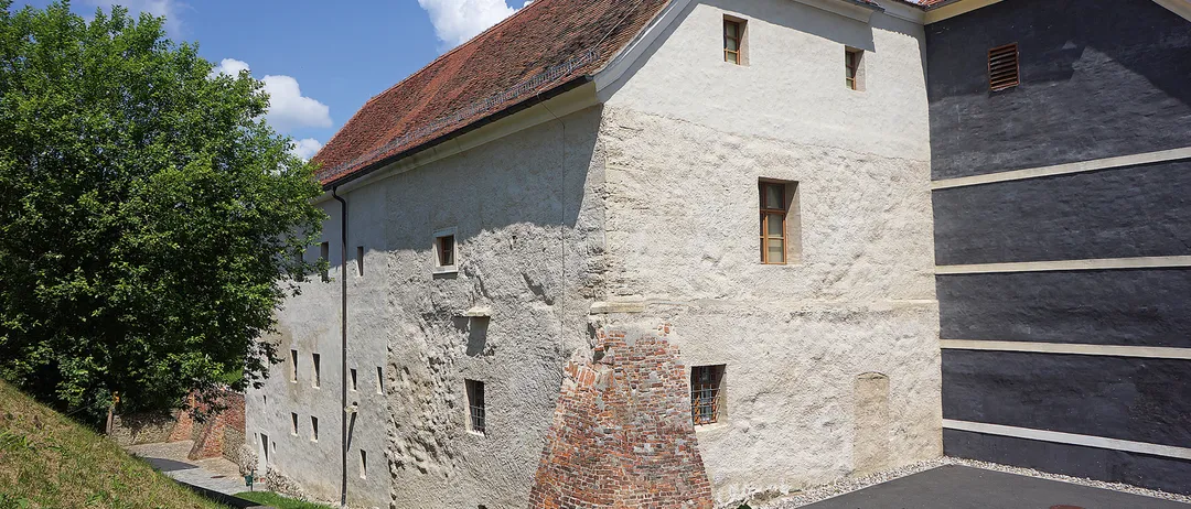 Ein altes Gebäude mit rotem Dach und weißen Wänden hat mehrere Fenster und einen Backsteinkamin. Das Gebäude ist von Grün umgeben, mit einem gepflasterten Weg, der zum Eingang führt.