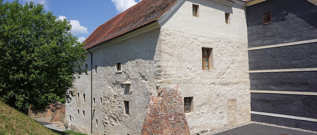 Ein altes Gebäude mit rotem Dach und weißen Wänden hat mehrere Fenster und einen Backsteinkamin. Das Gebäude ist von Grün umgeben, mit einem gepflasterten Weg, der zum Eingang führt.