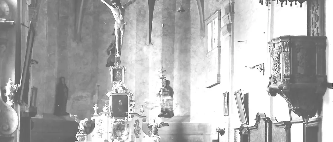 Schwarz-Weiß-Foto des Inneren einer Kirche. Ein Kruzifix mit einer Statue von Jesus hängt über einem Altar. Bänke sind in Reihen angeordnet und dem Altar zugewandt.