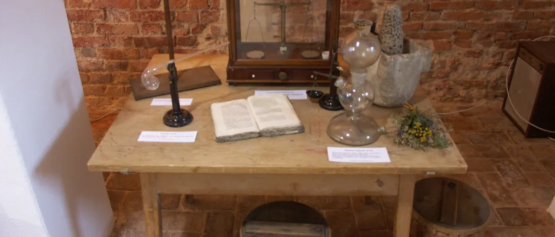 Ein hölzerner Tisch mit einer Waage, einem Mörser und verschiedenen Glasinstrumenten. Ein offenes Buch und ein Blumenstrauß sind ebenfalls auf dem Tisch. Der Tisch und die Wand bestehen aus Ziegelsteinen.