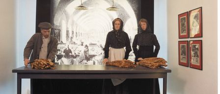 Three people are in a room with a table, one man and two women, all dressed in old-fashioned clothing. The table has a pile of leaves on it. Behind them is a large screen with a picture of a dining hall.