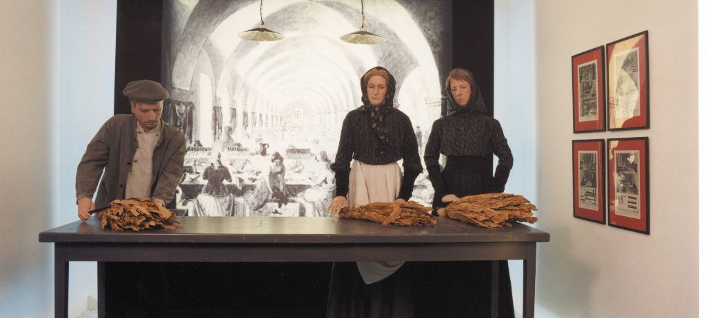 Drei Personen befinden sich in einem Raum mit einem Tisch, ein Mann und zwei Frauen, alle in altmodischer Kleidung gekleidet. Auf dem Tisch liegt ein Haufen Blätter. Hinter ihnen befindet sich eine große Leinwand mit einem Bild eines Speisesaals.