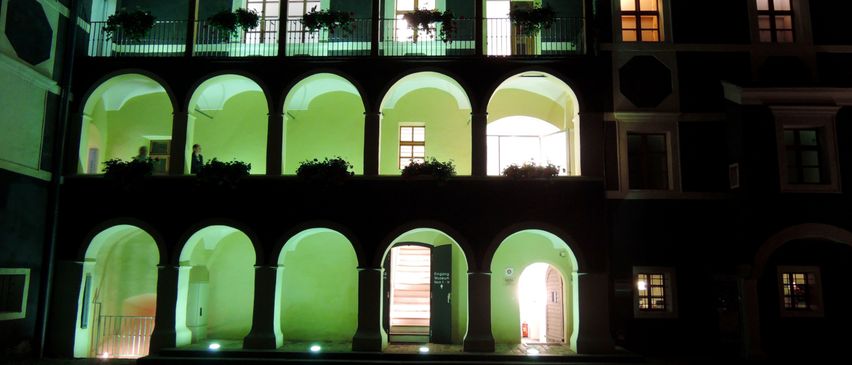 Ein Gebäude mit bogenförmigen Fenstern und Balkonen, die nachts mit grünem Licht beleuchtet sind. Zwei Personen stehen auf dem Balkon, und das Gebäude hat einen Ziegelboden.