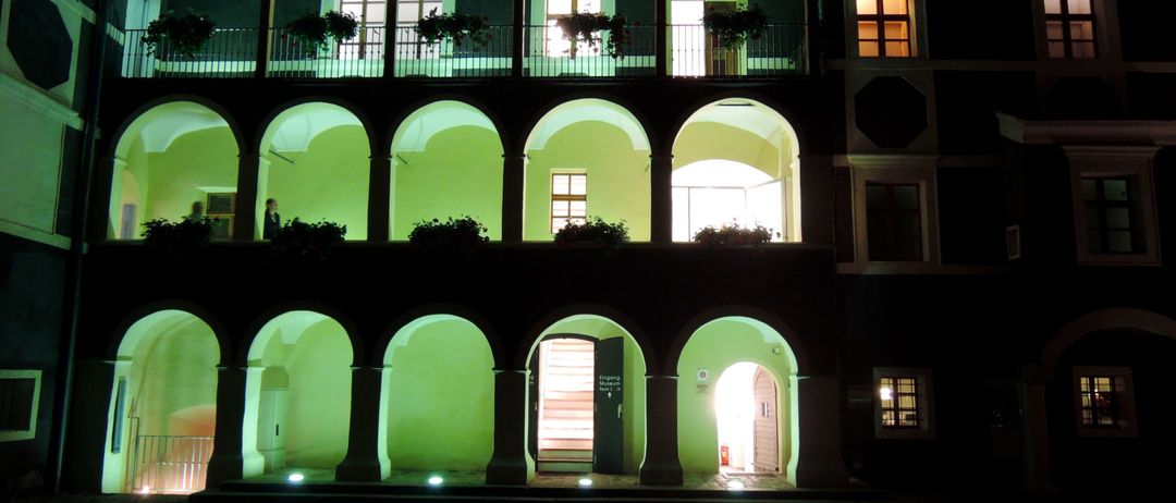 Ein Gebäude mit bogenförmigen Fenstern und Balkonen, die nachts mit grünem Licht beleuchtet sind. Zwei Personen stehen auf dem Balkon, und das Gebäude hat einen Ziegelboden.