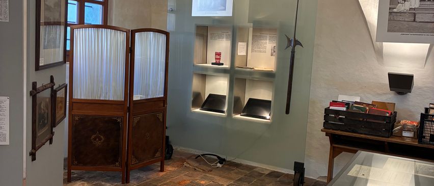 Ein Museumsraum zeigt Artefakte. Ein Vitrinenkasten mit einer Lanze befindet sich an der Wand. Ein Schreibtisch mit Büchern und eine große Vitrine sind im Raum.