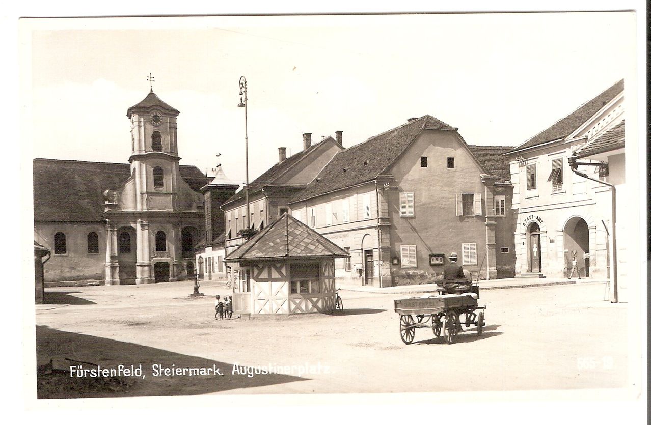 Ein altes Foto zeigt einen Stadtplatz mit einer Kirche, Gebäuden und einem Mann, der eine Kutsche zieht. Es stehen Menschen neben einem kleinen Gebäude.