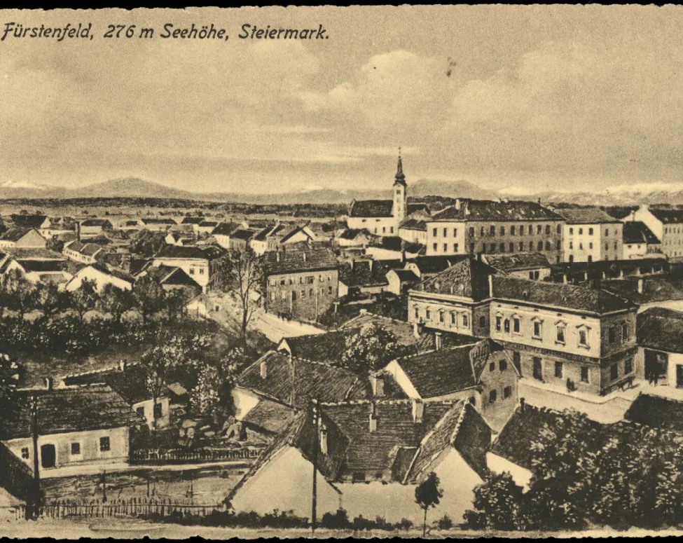 Ein altes Schwarz-Weiß-Foto von Furstenfeld, einer Stadt in der Steiermark, Österreich. Das Bild zeigt eine Panoramaansicht der Stadt mit mehreren Gebäuden, einem Turm und einer Kirche.