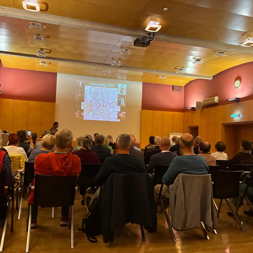 Eine Gruppe von Menschen sitzt in Stühlen in einem Raum und schaut sich eine Präsentation auf einer Projektionsleinwand an.