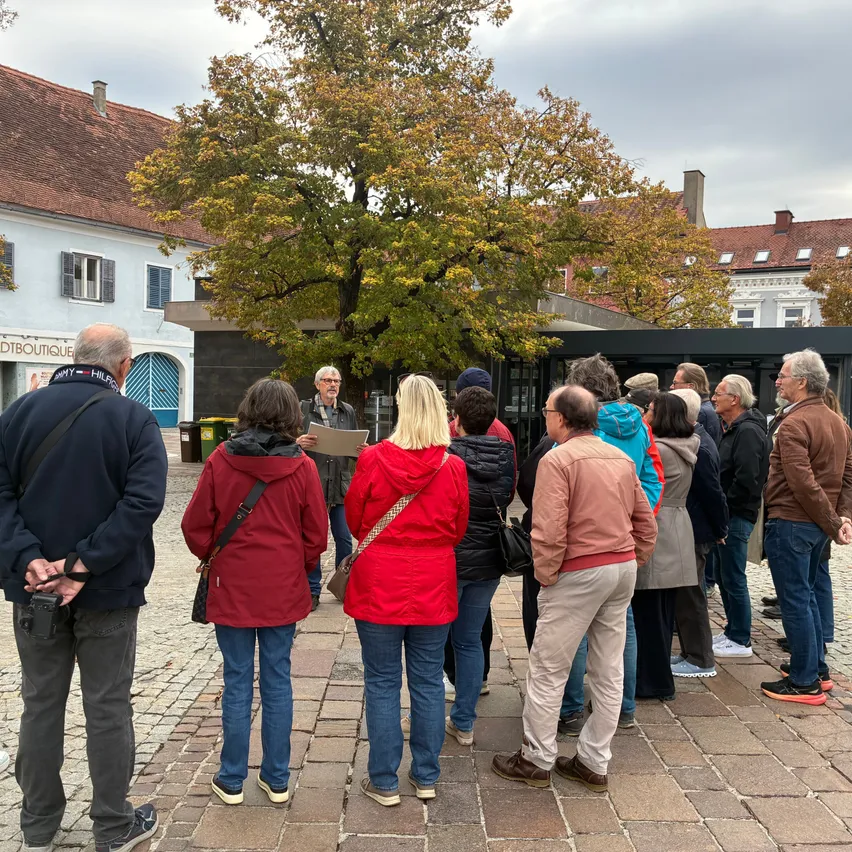 Eine Gruppe von Menschen versammelt sich auf einem Stadtplatz, wobei eine Person vor einem Baum steht, möglicherweise eine Rede hält. Die Gruppe ist in Winterkleidung gekleidet, und im Hintergrund befinden sich Gebäude.