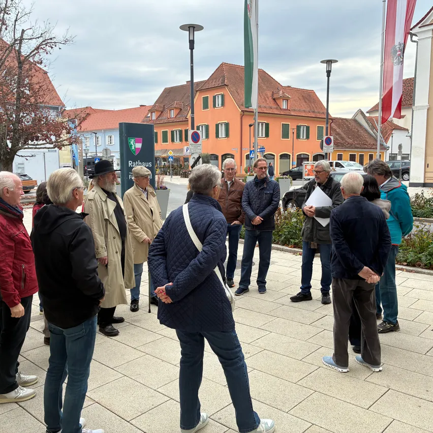 Eine Gruppe von Menschen steht draußen auf einem Bürgersteig. Einige tragen Jacken und Mützen. Ein Mann in einer blauen Jacke schaut auf ein Stück Papier.