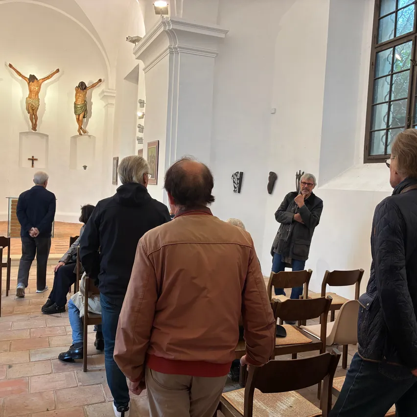 Eine Gruppe von Menschen versammelt sich in einer Kirche, einige stehen und andere sitzen auf Stühlen. Ein Mann steht vor einem Podium, und es gibt drei Statuen von Jesus an der Wand.