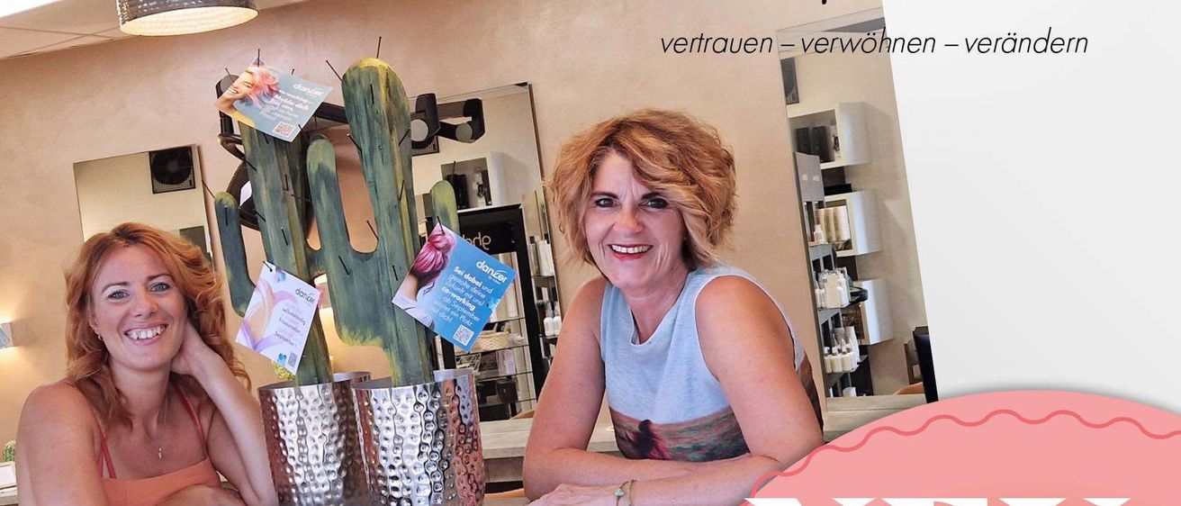 Zwei Frauen lächeln hinter einem Schreibtisch in einem Salon mit einem Kaktus in einem silbernen Topf, Lampen hängen von der Decke und ein Schild steht: 'Zusammen sind wir einfach besser!'