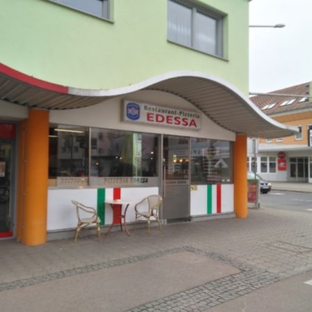 Ein Außenansicht eines Restaurants namens Edessa. Es hat ein weißes und grünes Farbschema, mit einem gebogenen Dach und zwei Stühlen und einem Tisch draußen. Es gibt auch ein Stoppschild und ein geparktes Auto auf der Straße.