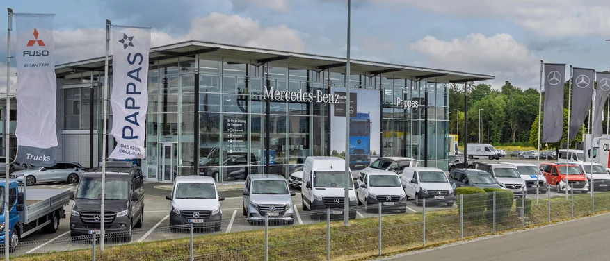 Ein Mercedes-Benz-Händler zeigt verschiedene Autos und Lastwagen auf seinem Parkplatz. Das Gebäude hat Glaswände und zeigt mehrere Banner.