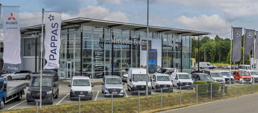 Ein Mercedes-Benz-Händler zeigt verschiedene Autos und Lastwagen auf seinem Parkplatz. Das Gebäude hat Glaswände und zeigt mehrere Banner.