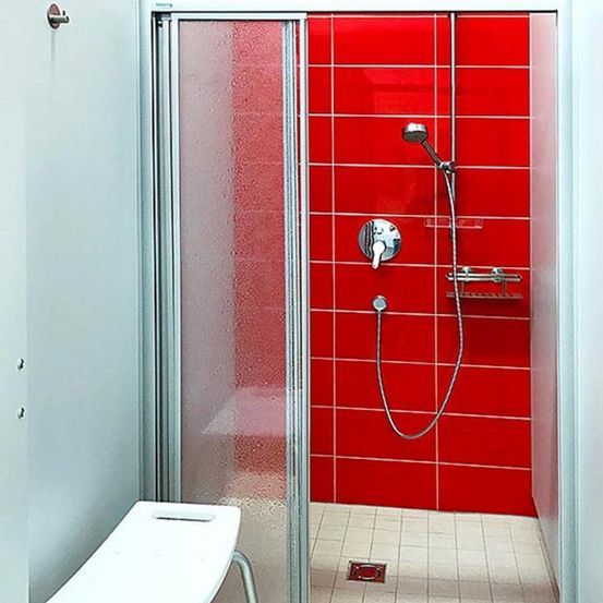 Eine moderne Duschkabine mit roter Fliesenwand, Glastür und Duschkopf mit Schlauch.