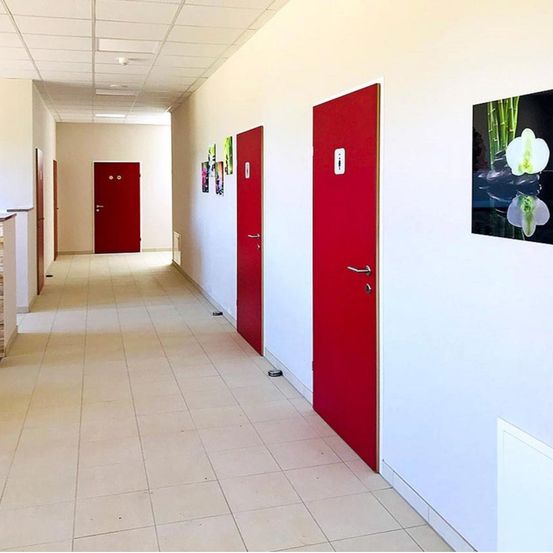 Ein leerer Flur mit weißen Fliesen und drei roten Türen mit Griffen und Türschildern.