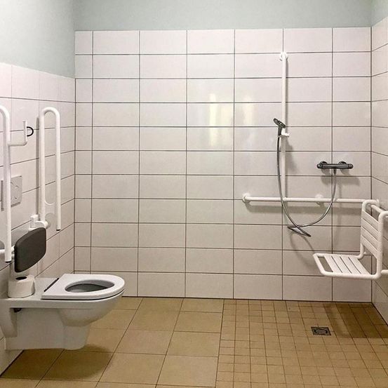 Ein weißes, sauberes Badezimmer mit Toilette, Dusche und weißem Fliesenboden. Es gibt eine Duschbrause, einen Duschständer und einen Handtuchhalter.