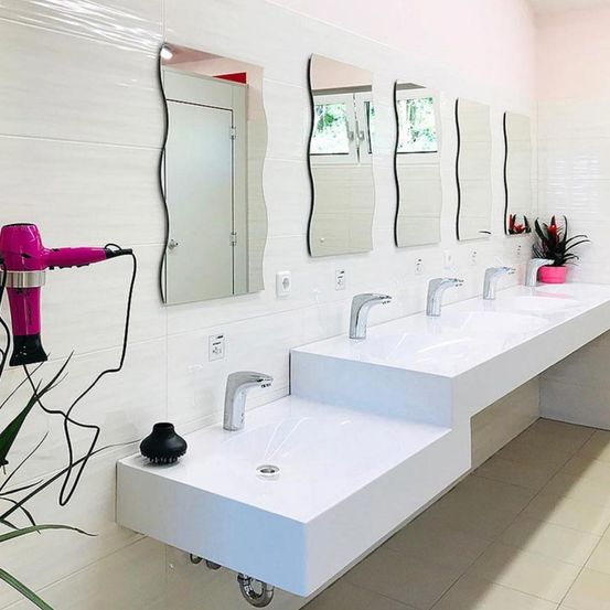 Ein rosa Föhn ist neben einem weißen Waschbecken mit Wasserhahn an der Wand montiert. Ein Topf mit einer Pflanze in Rosa steht neben dem Wandspiegel.