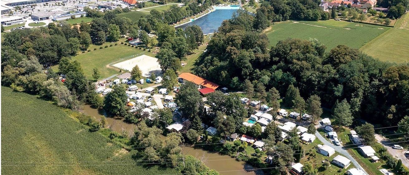 Luftaufnahme eines Campingplatzes am Fluss mit Schwimmbad, Gebäuden und Bäumen bei klarem blauem Himmel.