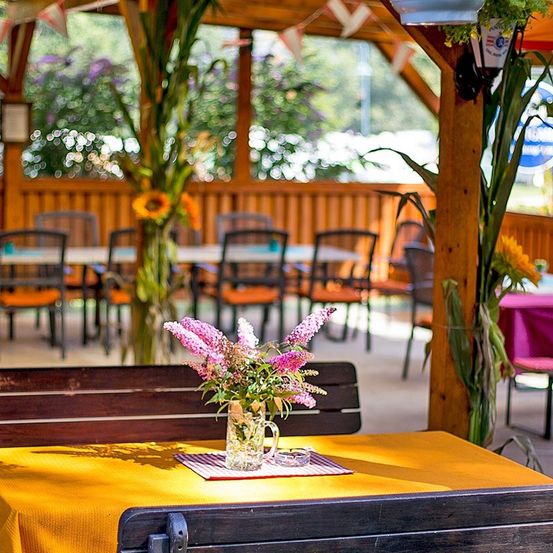 Ein Holztisch mit gelber Tischdecke und einem Blumenvase in einem Restaurant. Hinter dem Tisch sind weitere Tische und Stühle.