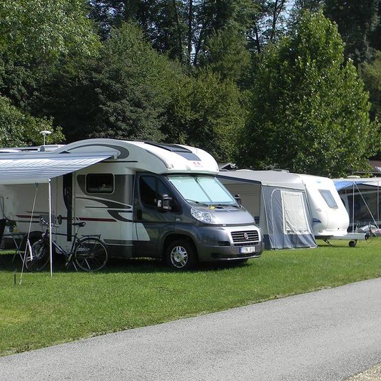 Ein grauer Wohnmobil, das auf einem Rasen neben einem Fahrrad und einem Tisch geparkt ist, mit anderen Wohnmobilen im Hintergrund unter Bäumen.
