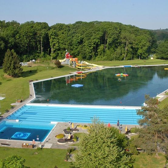 Luftaufnahme eines großen Freibades mit mehreren Bahnen, Rutschen und bunten aufblasbaren Spielzeugen im Wasser, umgeben von üppigem grünem Baumbestand und Rasen.