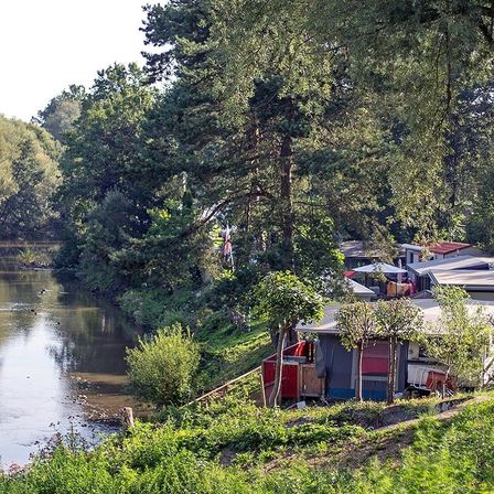 Eine Reihe von Campingzelten steht am Ufer eines Flusses, umgeben von Bäumen und üppiger Vegetation. Die Zelte sind auf einem grasbewachsenen Hügel aufgestellt, und der Fluss hat eine reflektierende Oberfläche mit einigen Wellen.