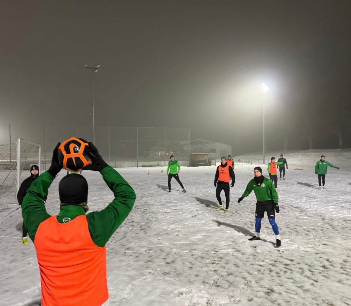Eine Gruppe von Fußballspielern trainiert bei Nacht auf einem verschneiten Feld, wobei ein Spieler einen Fußball hält.