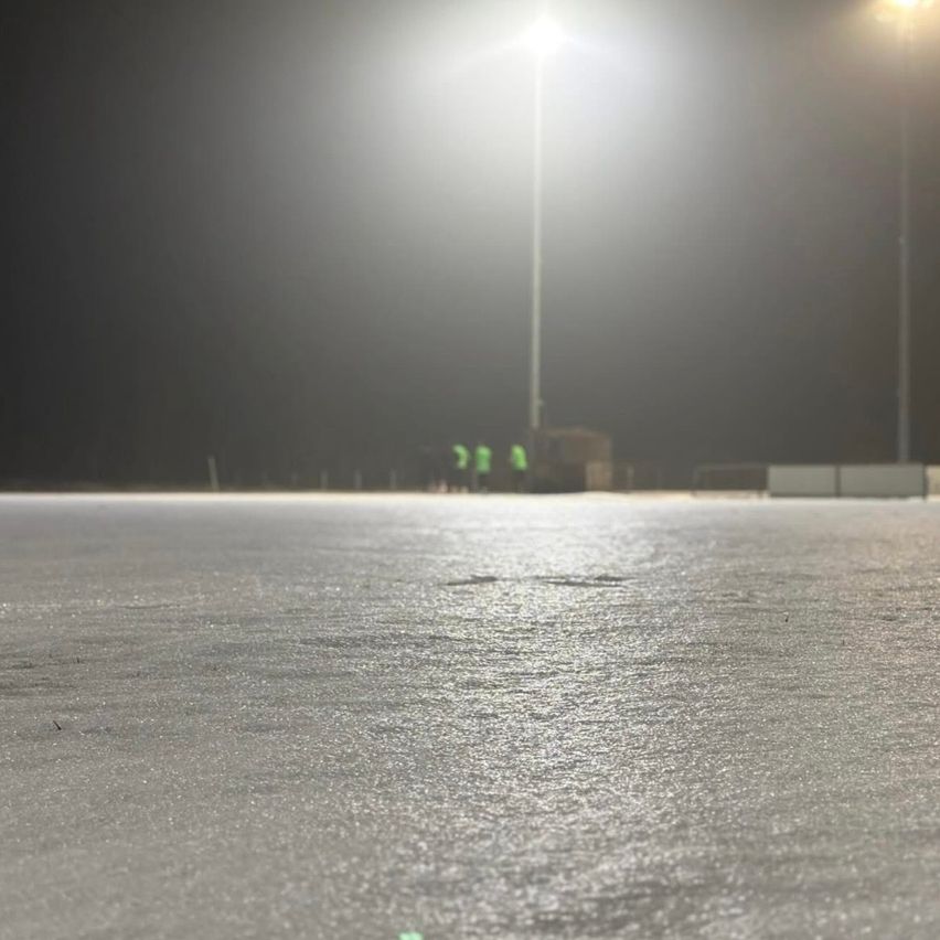 Eine nächtliche Ansicht einer leeren Eisbahn unter hellen Lichtern. Im Hintergrund stehen zwei Personen in grünen Westen.
