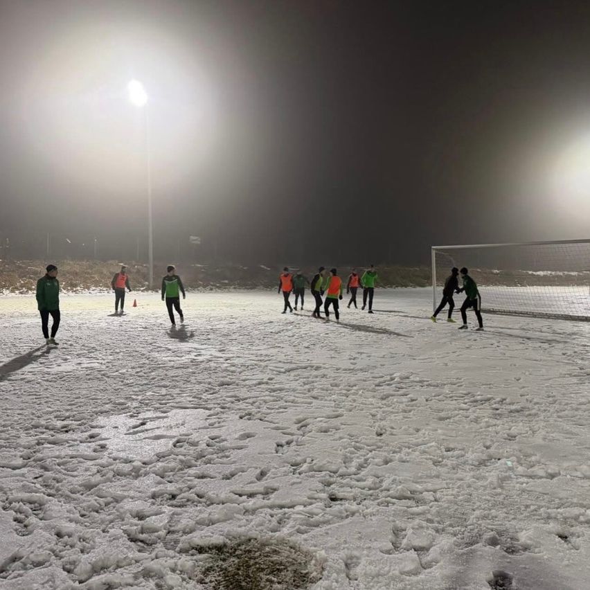Ein nächtliches Fußballtraining auf einem verschneiten Feld mit Spielern in grünen und orangefarbenen Trikots, beleuchtet von Flutlichtern.