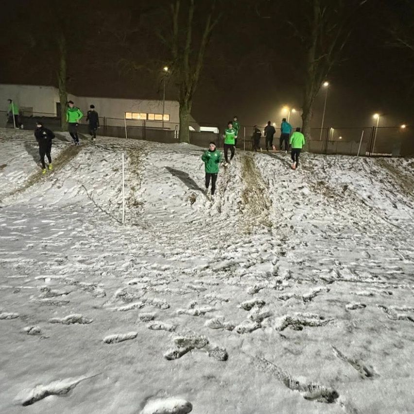 Eine Gruppe von Menschen in Grün joggt bei Nacht auf einem verschneiten Hügel. Im Hintergrund sind Bäume und Straßenlaternen zu sehen.