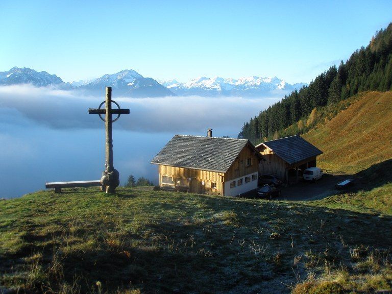 Ein Holzhaus mit grauem Dach steht auf einem Grashügel mit einem Kreuz und einer Bank. Autos sind davor geparkt. Nebel bedeckt das Tal mit schneebedeckten Bergen in der Ferne.