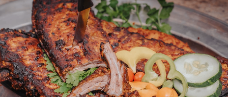 A plate with barbecued ribs, garnished with parsley, and a knife cutting into them. Accompaniments include sliced cucumbers, bell peppers, and tomatoes.