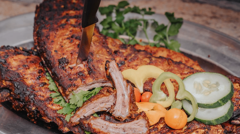 Ein Teller mit gegrillten Rippchen, garniert mit Petersilie, und einem Messer, das hineinschneidet. Beilagen sind geschnittene Gurken, Paprika und Tomaten.