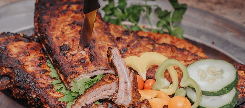 A platter of charred ribs is on a plate with a knife, accompanied by sliced cucumbers, bell peppers, and a sprig of herbs.