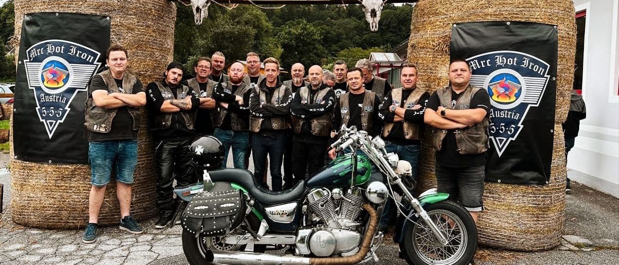Eine Gruppe von Motorradfahrern posiert mit ihrem Motorrad unter einem rustikalen Bogen. Das Schild darüber lautet 'MRC HOT IRON Bad Blumau', mit Kuhschädeln und Reifen geschmückt. Sie sind in Lederwesten gekleidet und einige tragen Uhren.
