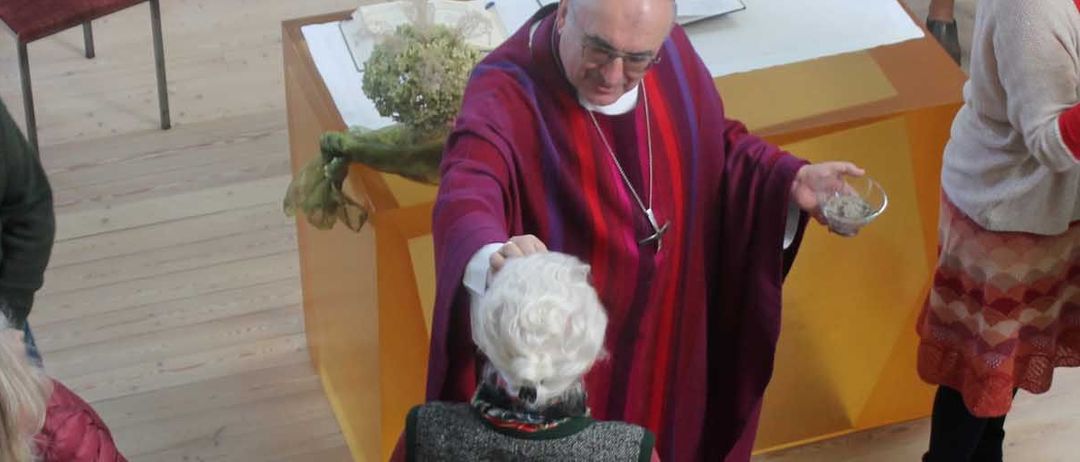 Ein Priester hält eine Schüssel und reicht eine ältere Frau. Die Leute sind um sie herum versammelt. Eine andere Person spielt Gitarre auf einem Stuhl. Ein Tisch mit Blumen steht vor ihnen.