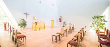 Eine leere Kapelle mit Holzboden, Reihen von Stühlen, einem gelben Altar mit Kerzen und einem Kreuz und einer Statue an der Wand.
