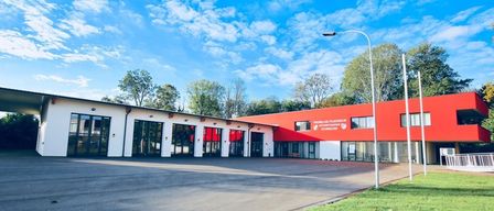 Ein Feuerwehrhaus mit roter Außenfassade und Glastüren. Im Hintergrund sind Bäume und eine Straßenlampe zu sehen.