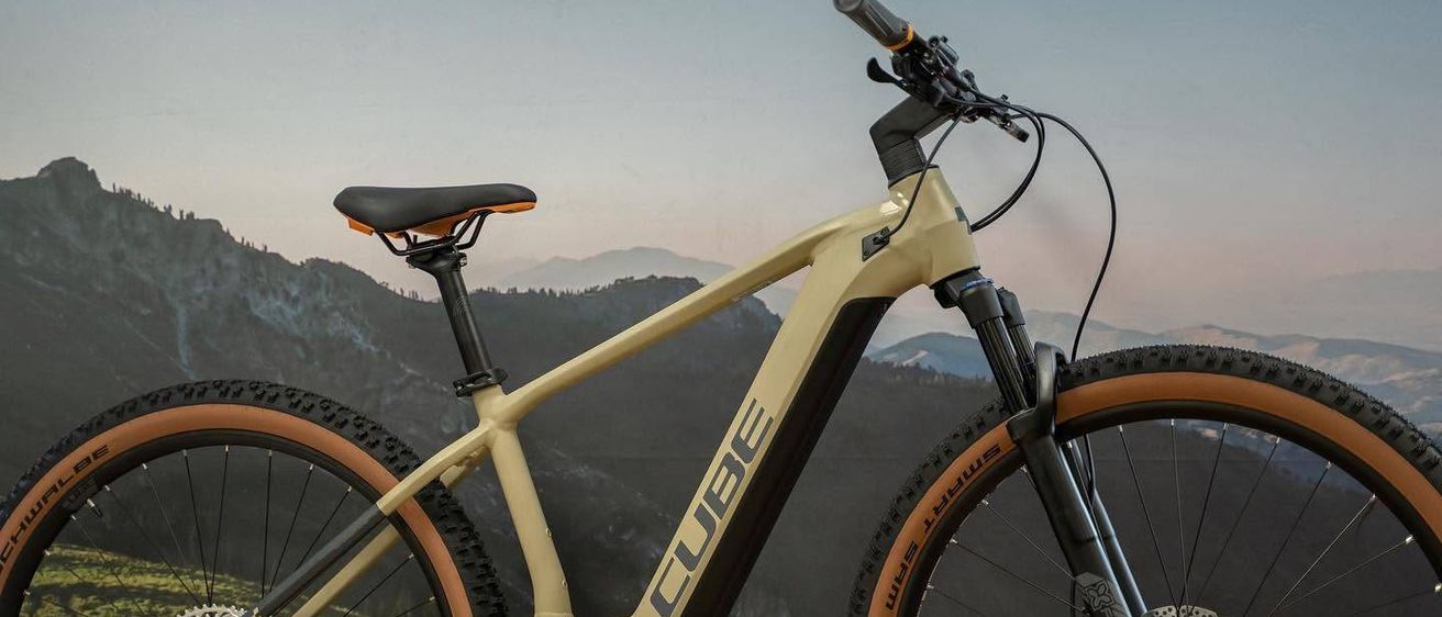 Ein beigefarbenes und schwarzes Elektro-Mountainbike mit 'CUBE'-Marke, ausgestellt auf einem Grashügel mit Gebirgshintergrund.