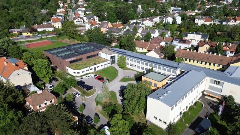 Luftaufnahme eines modernen Gebäudes mit Solarpanelen auf dem Dach, umgeben von Häusern und Bäumen. Mehrere Autos sind auf dem Parkplatz abgestellt.
