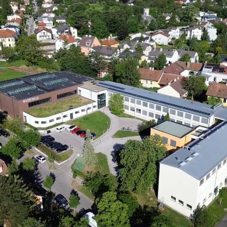 Luftaufnahme eines modernen Gebäudes mit Solarpanelen auf dem Dach, umgeben von Häusern und Bäumen. Mehrere Autos sind auf dem Parkplatz abgestellt.