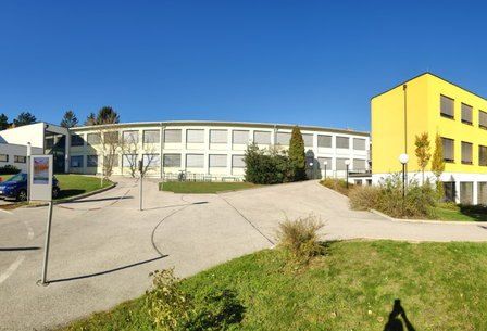 Ein modernes Gebäude mit gelber Vorderfassade befindet sich in einem Campus-Setting mit geparkten Autos davor, umgeben von Grünflächen und einem klaren Himmel.