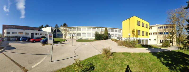 Ein modernes Gebäude mit gelber Vorderfassade befindet sich in einem Campus-Setting mit geparkten Autos davor, umgeben von Grünflächen und einem klaren Himmel.