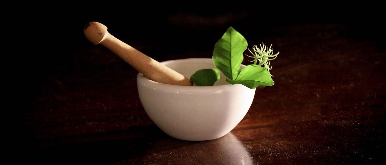 Ein weißer Mörser und Stößel mit einem grünen Blatt und kleinen Blüten auf einem dunklen Holztisch.