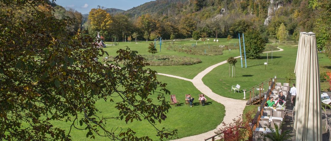 Ein Luftbild eines grünen Parks mit einem sich schlängelnden Pfad und Tischen und Stühlen an der Seite. In der Ferne befindet sich ein Gebirgszug mit Bäumen und Pflanzen.