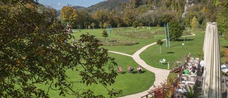 Ein Luftbild eines grünen Parks mit einem sich schlängelnden Pfad und Tischen und Stühlen an der Seite. In der Ferne befindet sich ein Gebirgszug mit Bäumen und Pflanzen.