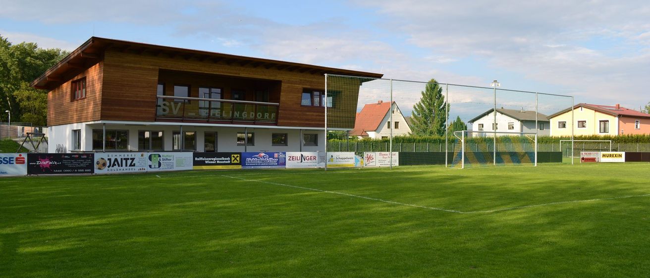 Ein Fußballfeld mit einem Gebäude im Hintergrund. Das Gebäude hat einen Balkon mit Glasgeländern und mehrere Banner an der Seite.