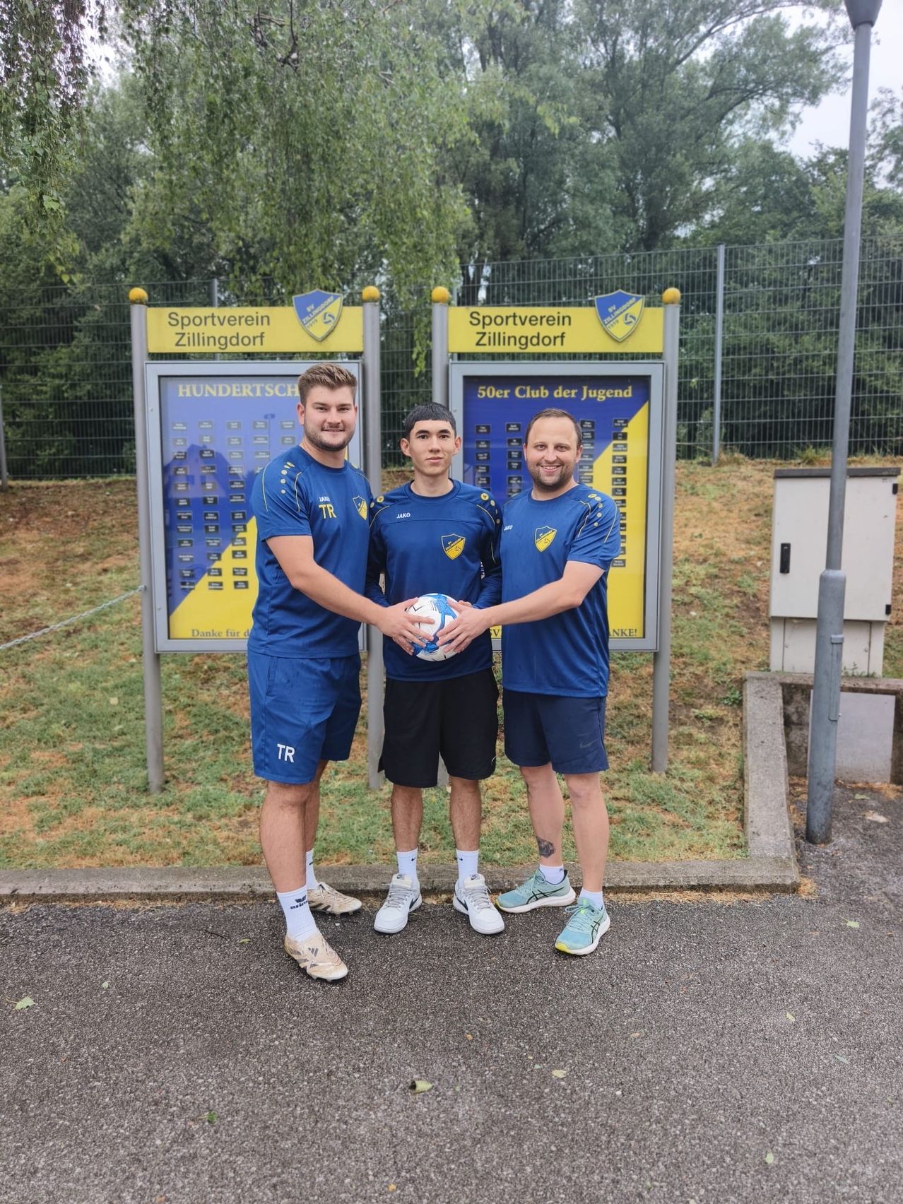 Drei Männer in blauen Uniformen stehen vor einem Schild des Sportvereins Zillingdorf und halten einen Fußball.